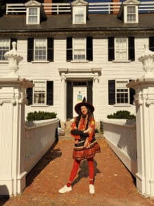 A woman dressed in a festive witch costume poses with her arms crossed in front of the Ropes Mansion in Salem, Massachusetts. The white historic house features black shutters, and she stands between two white gate pillars, smiling at the camera.