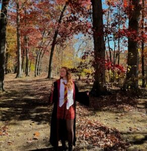 A person dressed in a Hogwarts-inspired costume, complete with a red and gold tie and robe, stands in a wooded area in Salem, Massachusetts. The scene is illuminated by sunlight filtering through vibrant autumn leaves, creating a magical and whimsical atmosphere.