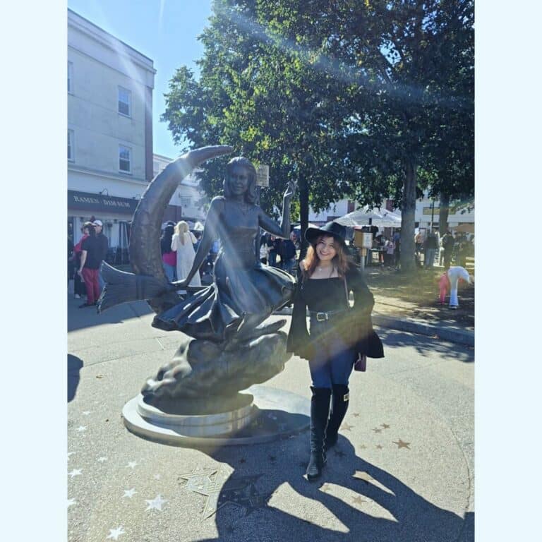 A woman in a black witch hat smiles while standing next to the "Bewitched" statue in downtown Salem. The bronze statue depicts the character Samantha Stephens sitting on a crescent moon with her broomstick, capturing her playful pose from the classic TV show "Bewitched." Sunlight filters through the trees, creating a warm, magical atmosphere.
