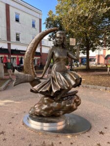 A statue of Samantha from the TV show "Bewitched" in Salem, Massachusetts. The statue features Samantha riding a broomstick, sitting on a crescent moon, with a flowing dress. The background includes a few shops, trees, and a clear blue sky.