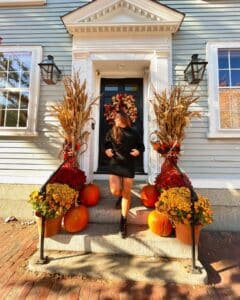 A woman stepping down the stairs of a house in Salem, Massachusetts, decorated with pumpkins, bright mums, cornstalks, and a colorful fall wreath, basking in warm sunlight.