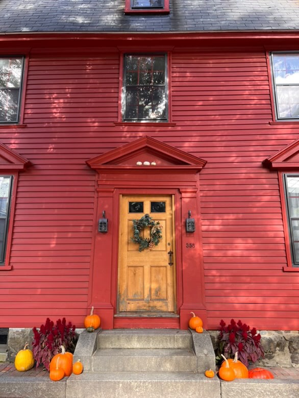 A red house in Salem, Massachusetts, featuring a wooden front door adorned with a wreath, steps decorated with pumpkins, and plants on either side.
