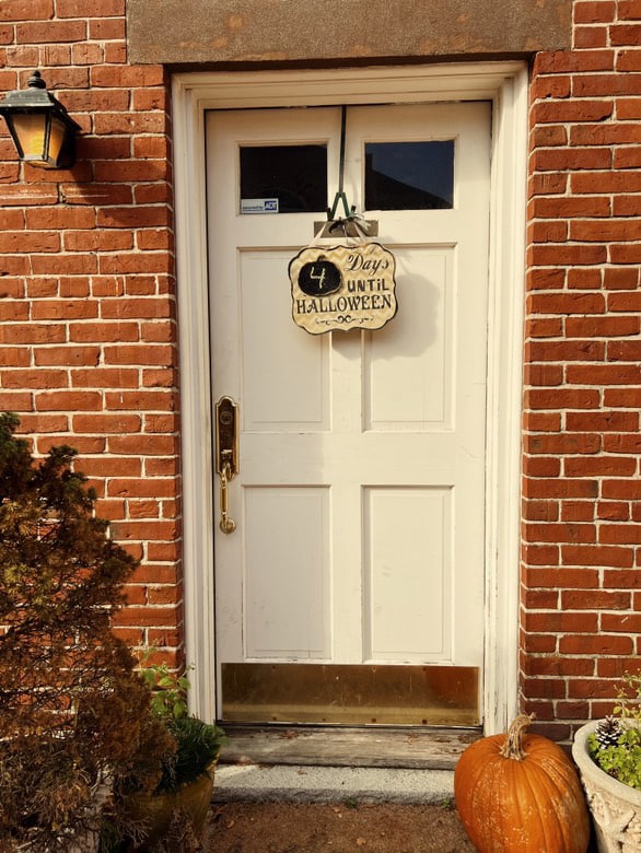 A white door in Salem, Massachusetts, with a hanging sign counting down "4 Days Until Halloween," surrounded by a red brick wall and autumn decorations like pumpkins.