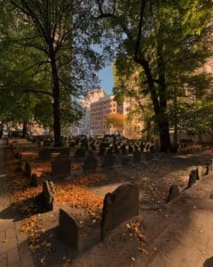 A daytime photo of the Old Burying Point Cemetery in Salem, Massachusetts, showing gravestones scattered throughout the area. Tall trees provide shade, and sunlight filters through the branches, illuminating fallen leaves on the ground. In the background, modern buildings rise above the cemetery, creating a mix of old and new.