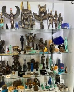 A display case in Salem showcases a collection of Egyptian-themed artifacts, including statues of deities, cats, and other symbolic figures. The items are crafted in various materials and colors, featuring intricate gold, bronze, turquoise, and lapis accents, creating a visually striking and mystical arrangement.