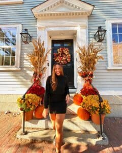 A woman walking down the steps of a beautifully decorated house in Salem, Massachusetts, surrounded by pumpkins, colorful mums, cornstalks, and a vibrant fall wreath on the door.