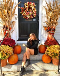 A woman sitting on the steps of a beautifully decorated house in Salem, surrounded by pumpkins, colorful mums, and festive cornstalks, with an autumn wreath hanging on the door.