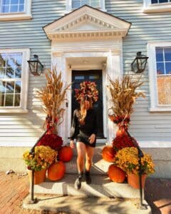 A woman stepping down the stairs of a house in Salem, Massachusetts, decorated with pumpkins, bright mums, cornstalks, and a colorful autumn wreath on the door.