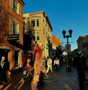 A lively scene on Essex Street in Salem, Massachusetts, featuring a colorful stilt performer dressed in a clown-like costume towering above the crowd. People in Halloween costumes and casual attire walk along the brick-lined street, surrounded by historic buildings bathed in warm sunlight.