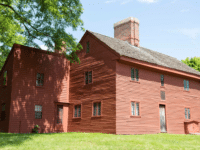 The Rebecca Nurse Homestead in Danvers, Massachusetts, showcasing a large red Colonial-style wooden house with a prominent brick chimney. The house is surrounded by a lush green lawn and shaded by tall trees, emphasizing its serene and historic setting. The bright blue sky adds to the picturesque view of this 17th-century homestead.