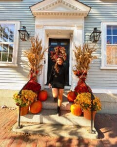 A woman standing on the steps of a colonial-style house in Salem, Massachusetts, decorated with pumpkins, autumn flowers, and cornstalks, capturing the festive spirit of fall.