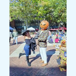 A lively scene in Salem shows a woman dressed in a stylish black outfit and wide-brimmed hat playfully posing with a man wearing a pumpkin head and a Halloween-themed suit. The backdrop features a bustling outdoor area with vendors, colorful decorations, and autumn foliage, capturing the festive spirit of the season.
