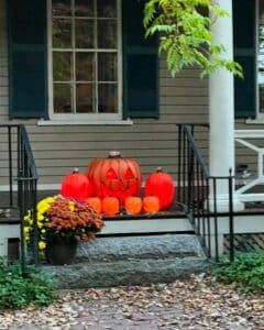 A front porch in Salem, Massachusetts, decorated with glowing jack-o'-lanterns of various sizes, surrounded by autumn leaves and a pot of colorful mums. The scene is framed by a classic colonial-style house with green shutters and white columns, creating a cozy Halloween atmosphere.