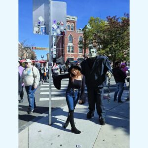 A woman dressed in a black witch hat and black outfit poses next to a tall Frankenstein's monster character on Essex Street in Salem, Massachusetts. The woman smiles playfully while the Frankenstein figure, holding a chain, stands beside her. In the background, people walk along the bustling street lined with historic buildings, enjoying the festive atmosphere.
