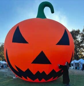 A giant inflatable jack-o'-lantern with a green stem stands on Salem Common, surrounded by festive booths and trees in the background, with a person dressed in a dark costume playfully posing next to it.