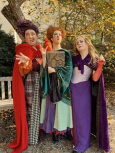 Three girls dressed as the Sanderson Sisters from the movie "Hocus Pocus" in Salem, Massachusetts. Each is wearing a colorful costume, with red, green, and purple capes, and making playful expressions while standing in front of fall foliage.