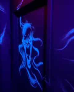 A glowing ghostly figure is painted on a dark wall, illuminated by blacklight in shades of eerie blue and purple. The artwork depicts a wailing spirit with sharp teeth and flowing, wispy tendrils, creating a haunting and atmospheric display.