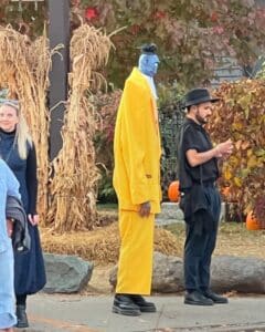 A tall person dressed in a vibrant yellow costume with blue face paint stands amidst a festive Halloween scene in Salem, Massachusetts. Surrounding them are cornstalk decorations, pumpkins, and people in various costumes, creating a lively autumn atmosphere.