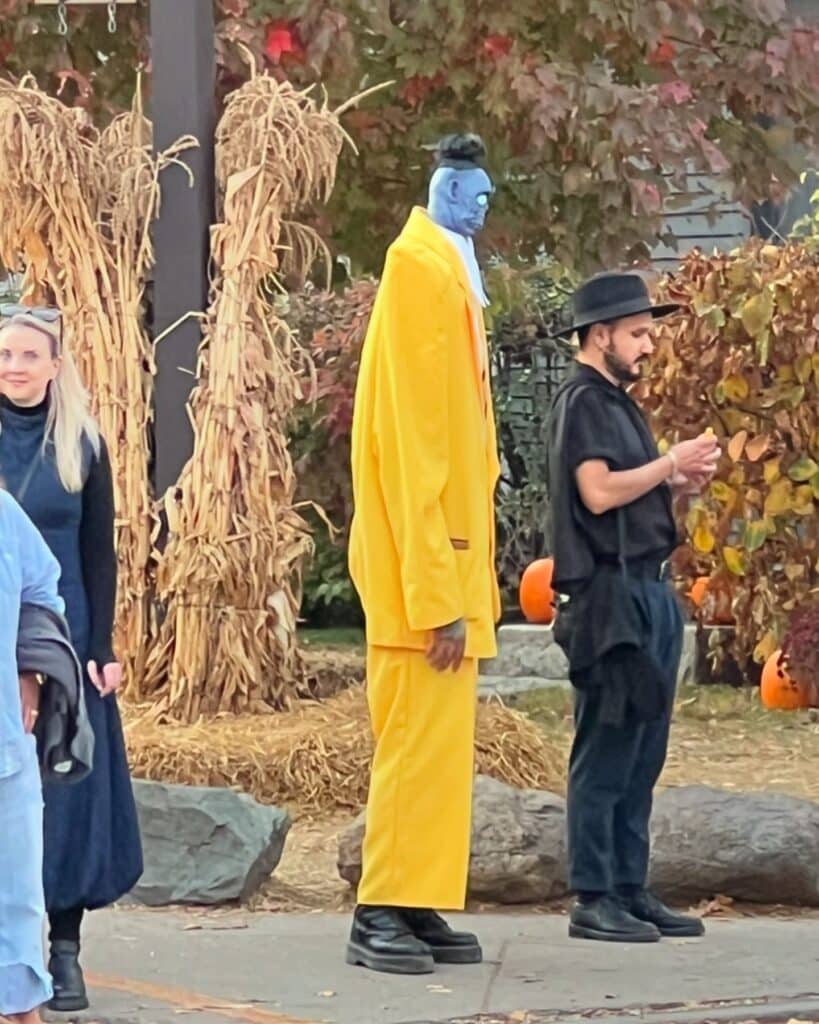 A tall person dressed in a vibrant yellow costume with blue face paint stands amidst a festive Halloween scene in Salem, Massachusetts. Surrounding them are cornstalk decorations, pumpkins, and people in various costumes, creating a lively autumn atmosphere.