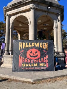 A Halloween Marketplace banner displayed in front of the stone bandstand at Salem Common in Salem, Massachusetts, featuring a jack-o'-lantern design and bold orange lettering.