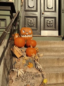 A collection of pumpkins, including one carved with a scary face, arranged on the steps of an old house in Salem, Massachusetts, featuring a dark, ornate door, burlap sacks, and fall decorations to create a festive Halloween atmosphere.