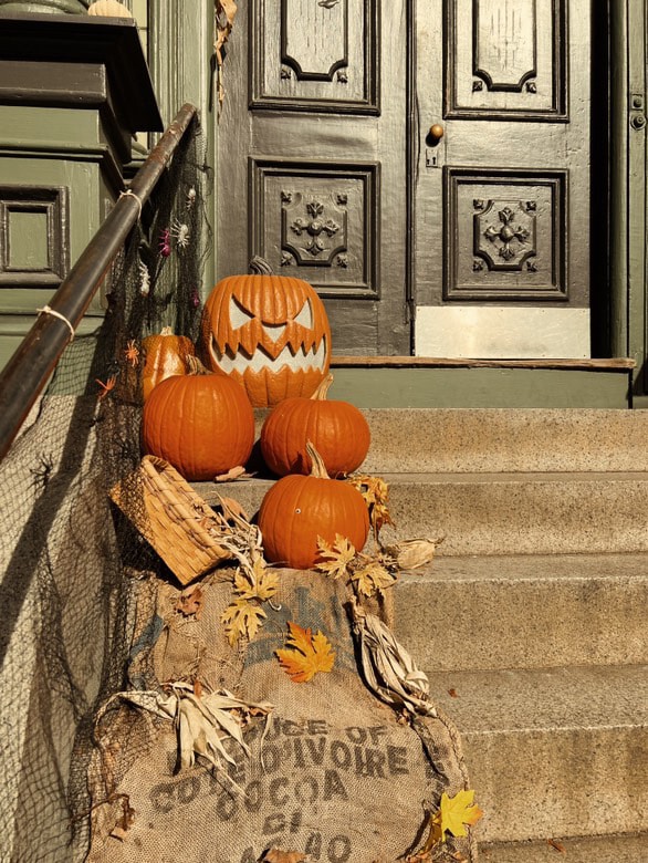 A collection of pumpkins, including one carved with a scary face, arranged on the steps of an old house in Salem, Massachusetts, featuring a dark, ornate door, burlap sacks, and fall decorations to create a festive Halloween atmosphere.