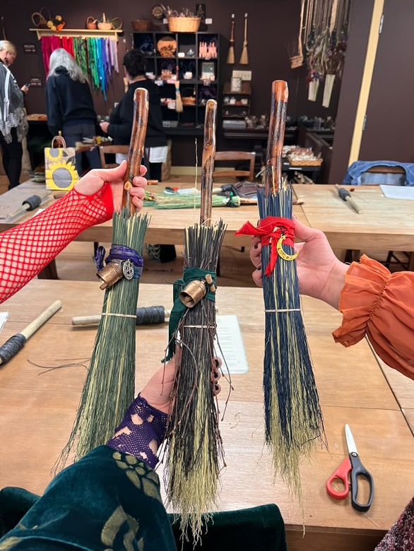 Three hands holding small handcrafted brooms decorated with colored ribbons and charms, displayed against a workshop setting. The ribbons are purple, green, and red, and the brooms have unique wooden handles. The background shows crafting supplies and shelves with materials.