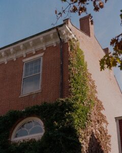 A photo of a historic brick building in Salem, Massachusetts, with ivy climbing up the side of the wall. The building has large windows with white trim and detailed decorative molding along the roofline. The blue sky and autumn leaves frame the scene.