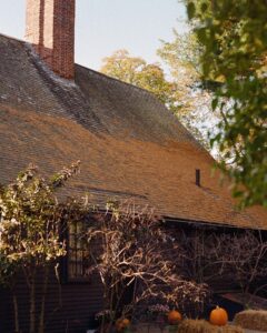 A photo of an old house in Salem, Massachusetts, with a sloped wooden shingle roof and a tall red brick chimney. The house has dark siding, surrounded by leafless branches and small trees. Pumpkins are scattered near the house, along with hay bales, giving it a classic autumn look.