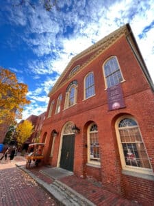 A photo of Salem's Old Town Hall, showing its red brick exterior, tall arched windows, and classic architecture. The sky above is partly cloudy, with a mix of blue and white, while colorful autumn leaves frame the scene.