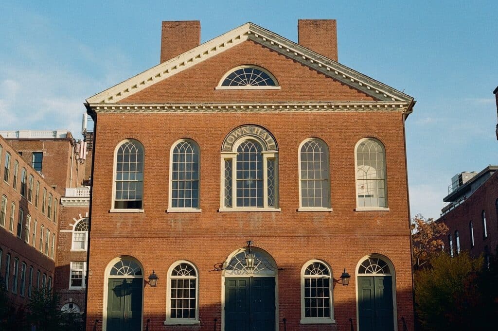 A photo of Salem's Old Town Hall, a historic red brick building with a symmetrical facade featuring arched windows and green double doors. The building's simple yet classic Federal-style architecture is highlighted by two tall chimneys and detailed trim along the roofline. The warm afternoon sunlight creates soft shadows, adding to the building's timeless charm.
