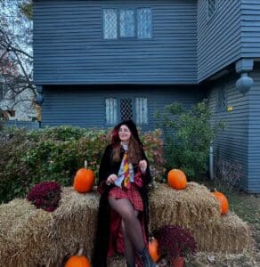 "A person dressed in a Halloween costume sitting on hay bales surrounded by pumpkins and flowers in front of the historic Witch House in Salem, Massachusetts, a dark timber-framed building with lattice windows and iconic colonial architecture.