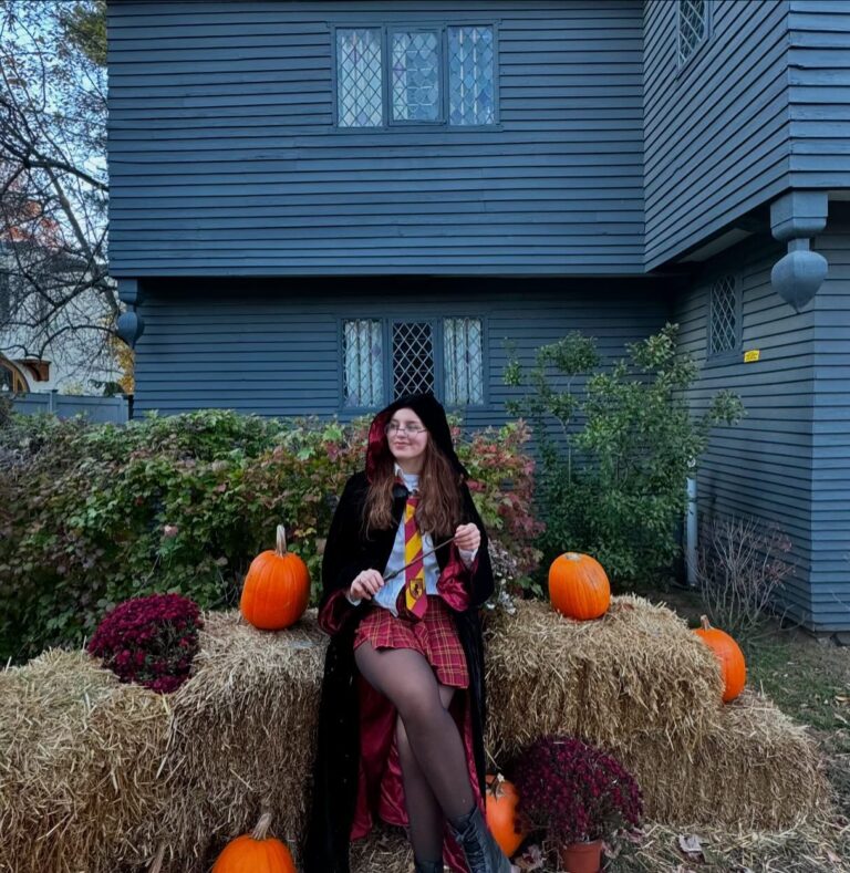 "A person dressed in a Halloween costume sitting on hay bales surrounded by pumpkins and flowers in front of the historic Witch House in Salem, Massachusetts, a dark timber-framed building with lattice windows and iconic colonial architecture.