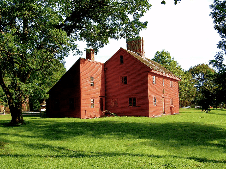 The Rebecca Nurse Homestead in Danvers, Massachusetts, features a red-painted wooden Colonial house with two large chimneys, surrounded by lush green grass and shaded by trees. This historic site offers a glimpse into life during the 1600s and is a key location tied to the Salem Witch Trials of 1692.