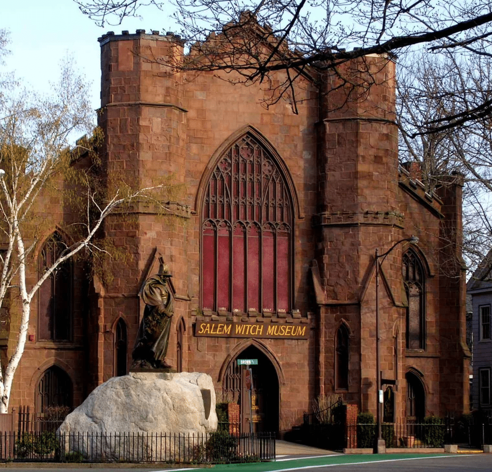 Exterior view of the Salem Witch Museum in Salem, Massachusetts. The historic building is constructed of red stone, with Gothic-style arched windows and an imposing presence. In front of the museum stands a statue of a witch on a large stone pedestal, symbolizing the town's connection to the infamous Salem Witch Trials. Bare trees surround the building, adding a stark, atmospheric touch to the scene.