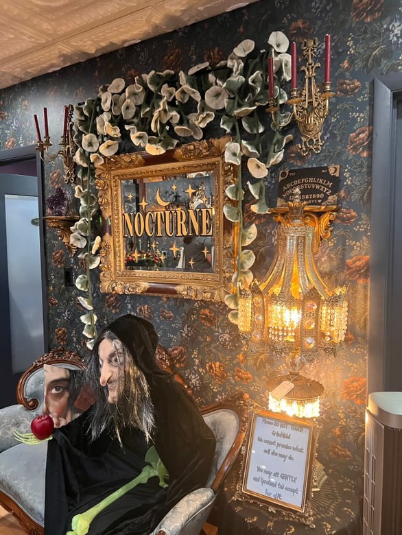 A witch-themed display inside the Nocturne shop in Salem, Massachusetts, featuring an ornate frame with the word "Nocturne" surrounded by faux flowers, a chandelier, and a mannequin dressed as a witch holding an apple.