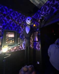 A life-sized figure of Jack Skellington from *The Nightmare Before Christmas* stands in a dimly lit room in Salem, Massachusetts. The figure is surrounded by gothic decor, including patterned wallpaper, eerie lighting, and glowing pumpkins at his feet, creating a spooky yet whimsical atmosphere.