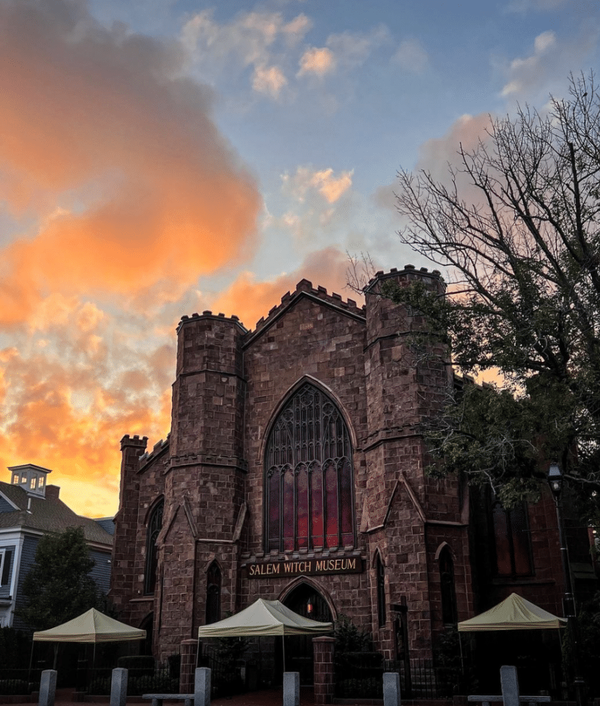 A striking image of the Salem Witch Museum at sunset, featuring its iconic Gothic-style architecture made of dark stone. The building’s tall arched windows glow with red light, while the sky behind is painted with vibrant orange and pink hues. Bare trees frame the scene, adding to the museum’s historic and mysterious atmosphere.