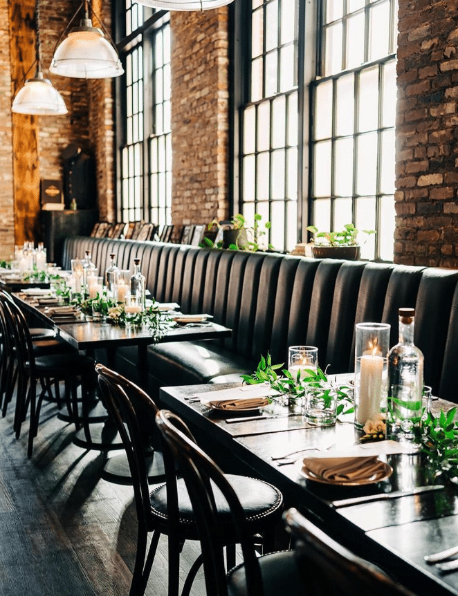 The dining area at Ledger Restaurant in Salem, Massachusetts, is elegantly arranged with black leather seating, dark wooden tables, and minimalist greenery accents. Soft candlelight and the industrial-chic ambiance created by exposed brick walls and large paneled windows provide a warm and inviting atmosphere, perfect for intimate gatherings or special celebrations.