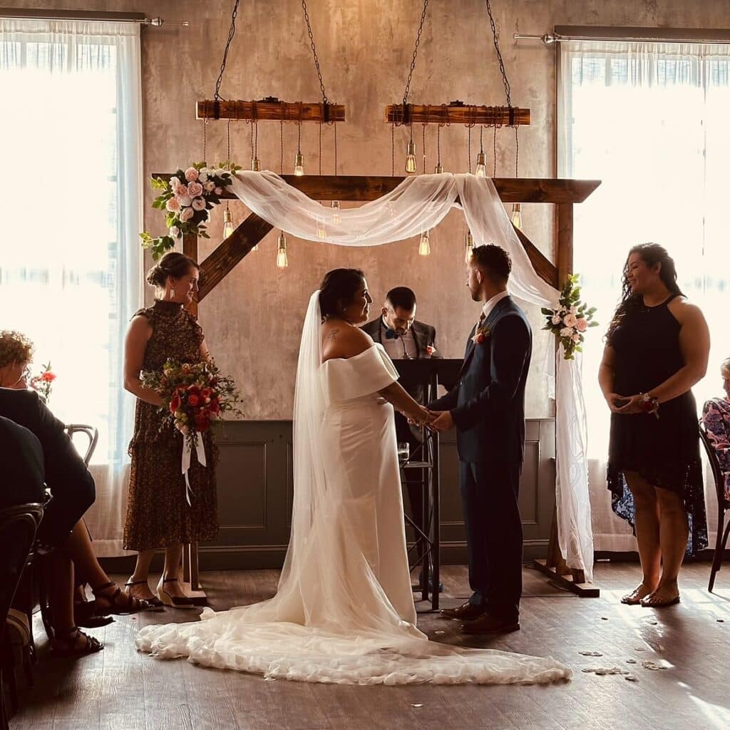 A beautiful wedding ceremony at Ledger Restaurant in Salem, Massachusetts, featuring an elegant rustic arch adorned with white drapery and floral arrangements. The intimate setting, with warm lighting and a historic ambiance, creates a romantic and memorable atmosphere for the couple and their guests.