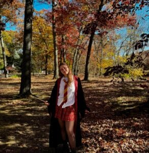A person dressed in a Hogwarts-themed costume, featuring a red and gold tie, plaid skirt, and robe, stands in a sunlit forest in Salem, Massachusetts. The surrounding trees display vibrant autumn foliage, with fallen leaves blanketing the ground, creating a warm, seasonal atmosphere.