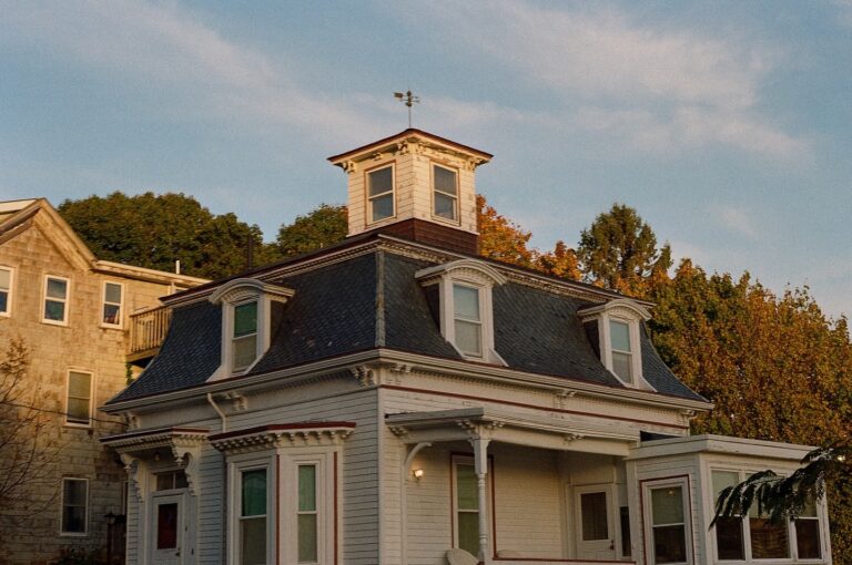 A photo of Max and Dani's house from the movie "Hocus Pocus" in Salem, Massachusetts. The house features a mansard roof with dormer windows, a small cupola at the top with a weather vane, and intricate trim details. The white siding and dark roof contrast against the surrounding fall foliage, adding to the house's charming, classic New England look.