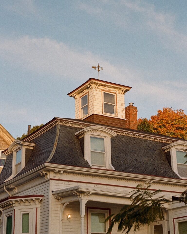 A photo of Max and Dani's House in Salem, Massachusetts, showing its distinctive architectural style with a mansard roof, dormer windows, and a small cupola on top. The house features white siding, detailed trim, and a weather vane on the cupola, with the soft light of evening giving it a warm and inviting appearance.