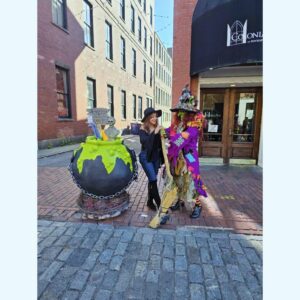A woman wearing a black witch hat smiles while posing next to Borah Brewington Snaggletooth XIII, a colorful street performer in Salem, Massachusetts. Borah is dressed in a vibrant, patchwork witch costume, holding a broom and standing beside a large cauldron decorated with bright green slime and Halloween props. The scene is set on a cobblestone street, with brick buildings in the background, capturing the whimsical, festive atmosphere of Salem.
