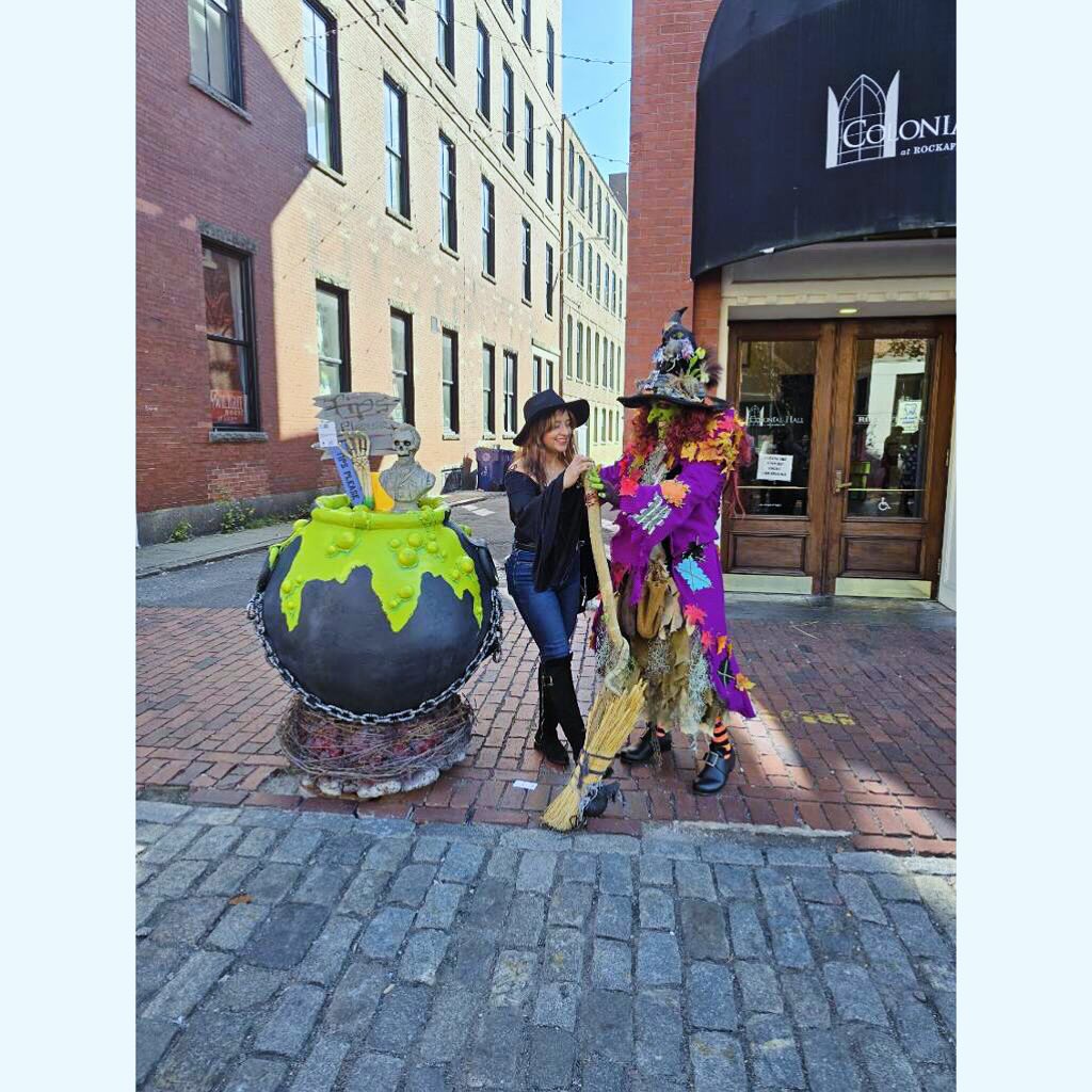A woman wearing a black witch hat smiles while posing next to Borah Brewington Snaggletooth XIII, a colorful street performer in Salem, Massachusetts. Borah is dressed in a vibrant, patchwork witch costume, holding a broom and standing beside a large cauldron decorated with bright green slime and Halloween props. The scene is set on a cobblestone street, with brick buildings in the background, capturing the whimsical, festive atmosphere of Salem.