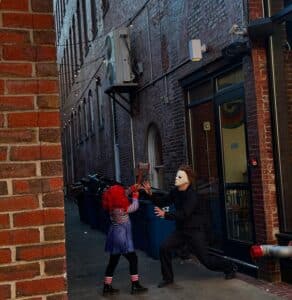 Two costumed individuals are playfully reenacting a dramatic scene in an alley off Essex Street in Salem, Massachusetts. The alley features red brick walls, string lights, and garbage bins, adding an urban backdrop to the spirited Halloween-themed moment.