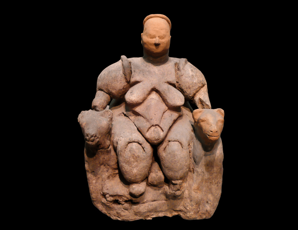Ancient figurine of a Mother Goddess, seated with a rounded body, symbolizing fertility and abundance. The goddess holds two large feline heads on either side, possibly representing her connection to animals or protective qualities. The figurine is crafted from clay or stone, with a simple, abstract facial expression, characteristic of early goddess sculptures