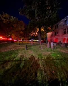 A nighttime photo of the Old Burying Point Cemetery in Salem, Massachusetts, showing gravestones scattered across the grassy area. Trees surround the cemetery, lit by eerie red and orange lights. A historic wooden building is visible to the right, adding to the spooky atmosphere.