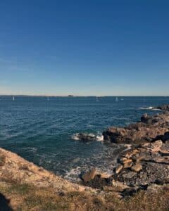A photo of the ocean in Salem, Massachusetts, showing calm blue waters under a bright blue sky. Small waves crash against the rocky shoreline in the foreground, while several sailboats are visible on the horizon.