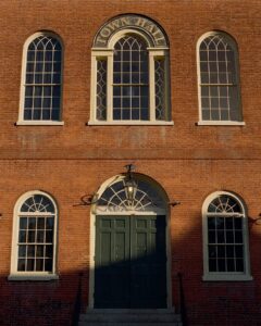 The front facade of the Old Town Hall in Salem, Massachusetts, featuring a red brick exterior with arched windows and a large green double door. Above the door, an arched sign reads "Town Hall," and sunlight casts soft shadows across the building, highlighting its historic architecture.